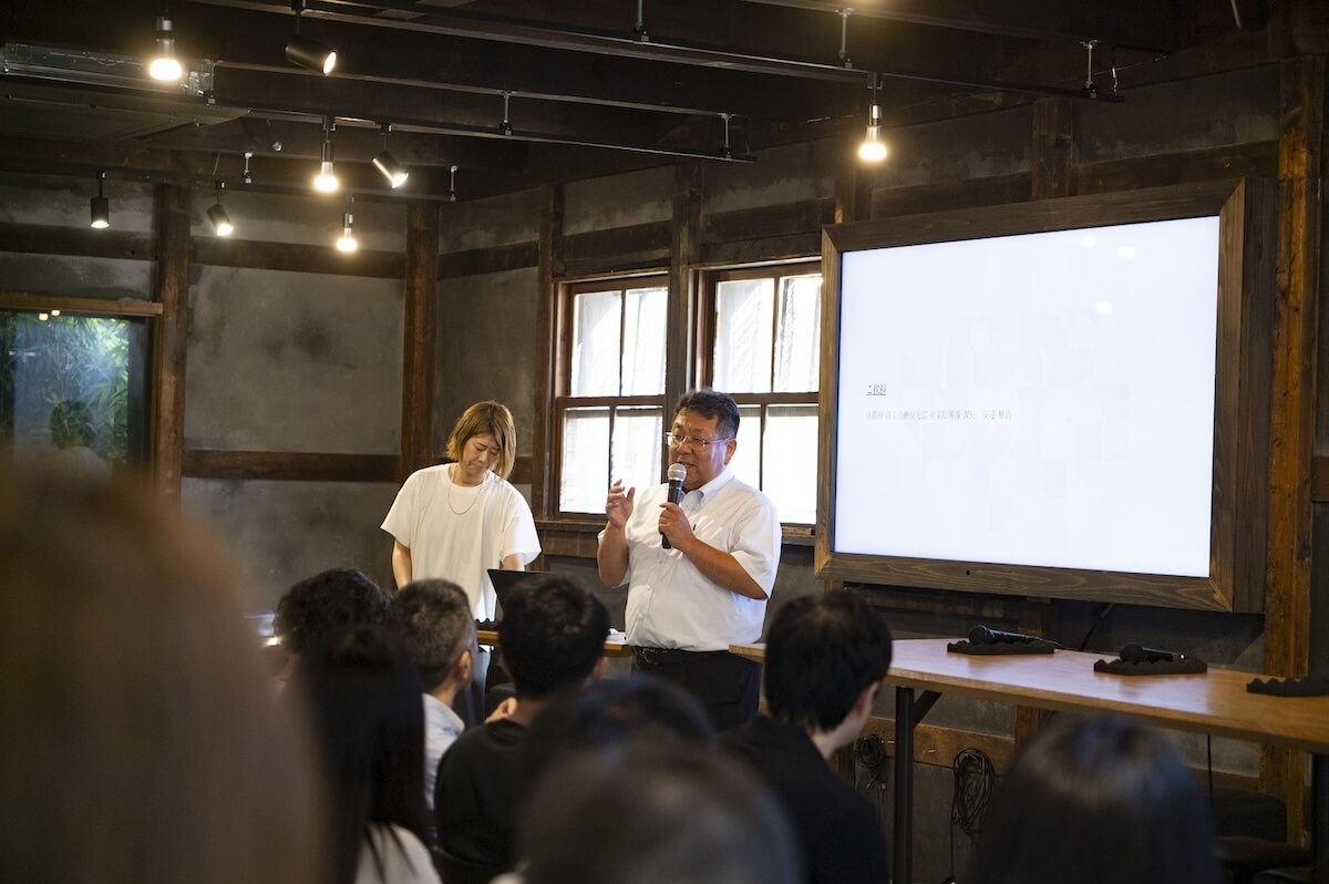 白シャツ姿の男性がマイクを持ち話している様子